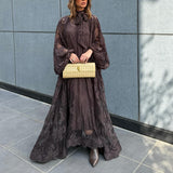 Woman in a long dark lace dress holding a gold clutch against a gray tiled wall.