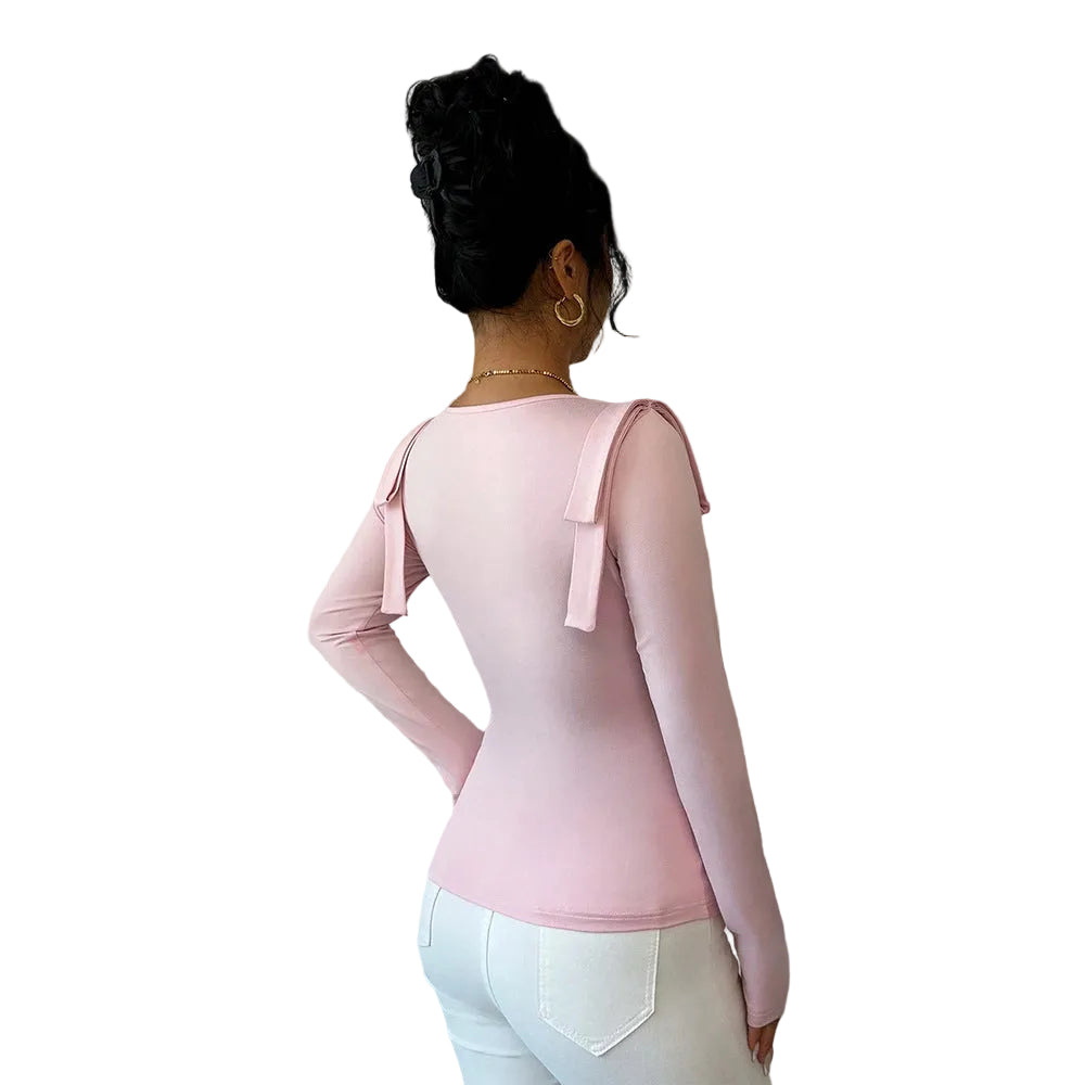 Woman wearing a pink long-sleeve top with bow details, standing in a room with curtains and a plant.