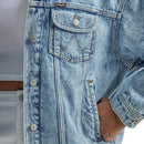 Close-up of a light blue Wrangler denim jacket worn over a white shirt, showing the chest pocket with a silver button, contrast stitching, and the Wrangler logo tab.